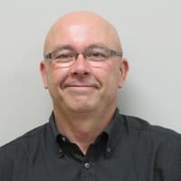 Smiling bald man with glasses wearing a black button-down shirt against a plain background.